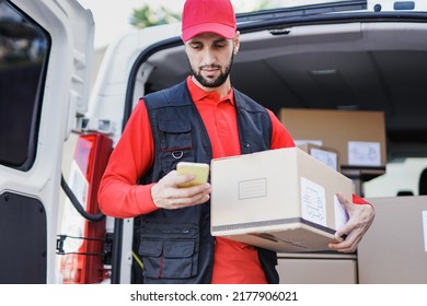 Young caucasian courier man using mobile phone while holding delivery box out of van - Powered by Shutterstock