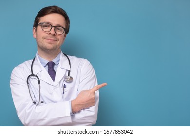 Young caucasian doctor pointing up showing on copyspace smiling and looking at camera confident. Studio shot on blue wall