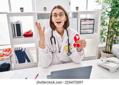 Young caucasian doctor woman holding support red ribbon celebrating victory with happy smile and winner expression with raised hands  - Shutterstock ID 2256572697