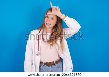 Young caucasian doctor woman wearing medical uniform and stethoscope over blue background making fun of people with fingers on forehead doing loser gesture mocking and insulting.