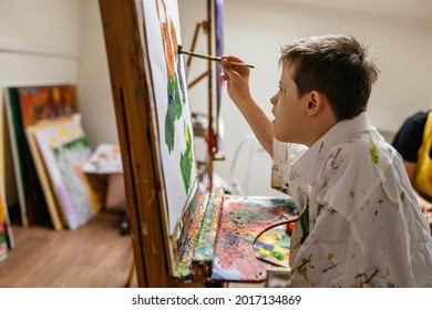 Young Caucasian Down Syndrome Boy Sitting At Chair With Colors And Painting On Easel With Concentrated And Calm Painting. Concept For Education For Disabled Kid.