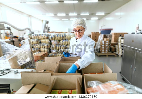 Young Caucasian employee in\
sterile uniform using tablet for logistic. Food factory\
interior.