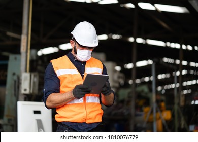 Young Caucasian Engineering Man Worker Working On Digital Tablet Computer At Manufacturing. Worker Man Wearing Face Mask Prevent Covid-19 Virus And Protective Hard Hat. 