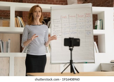 Young Caucasian English Teacher Make Flip Chart Presentation Record Video On Cellphone. Female Tutor Or Coach Present New Topic On Whiteboard, Talk On Webcam Live Broadcast Or Event On Smartphone.