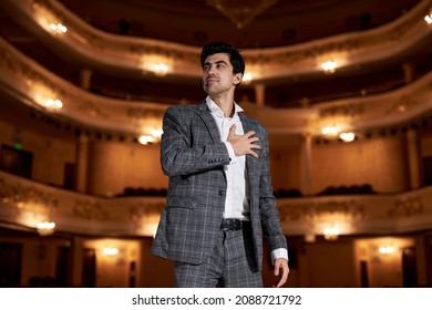 Young Caucasian European Actor Performing A Monologue In Theater To Audience, Talented Brunette Guy Portrait In Gray Elegant Formal Suit, Standing On Stage Alone, Black Background. Live Performance