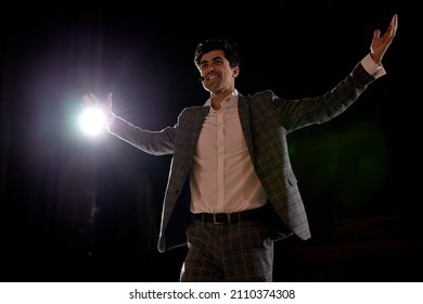 Young Caucasian European Actor Performing A Monologue In Theater To Audience, Talented Brunette Guy Portrait In Gray Elegant Formal Suit, Standing On Stage Alone, Black Background. Live Performance