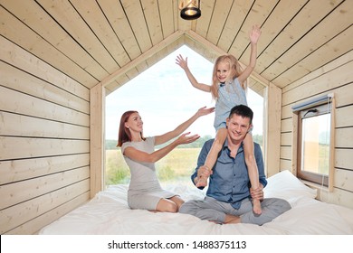 Young Caucasian Family Couple With Baby Daughter In A Small Modern Rustic House With A Large Window. Lying On The Bed, Hugging, Playing And Looking Out The Window. Weekend Vacation Concept Outside The