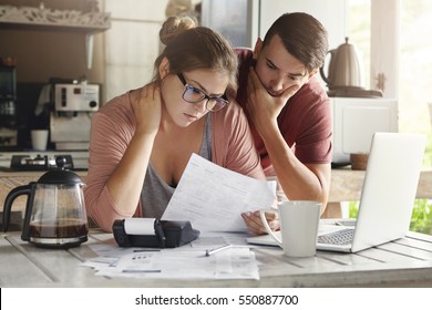 Young Caucasian family having debt problems, not able to pay out their loan. Female in glasses and brunette man studying paper form bank while managing domestic budget together in kitchen interior