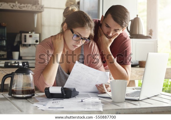 Young Caucasian family having debt problems, not\
able to pay out their loan. Female in glasses and brunette man\
studying paper form bank while managing domestic budget together in\
kitchen interior