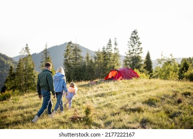 Young Caucasian Family With Little Girl Walk Together On Green Meadow While Traveling With Tent In The Mountains During Sunset. Happy Family Spending Summer Vacation At Campsite