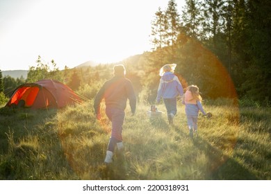 Young Caucasian Family With Little Girl Walk Together On Green Meadow While Traveling With Tent In The Mountains During Sunset. View From The Backside. Image With Lens Flare