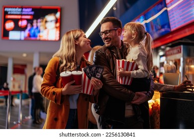 A young Caucasian family is at the movie theater, bonding as they're ready to see a movie together. The father is carrying their child and the mother is carrying popcorn, tickets and beverages.