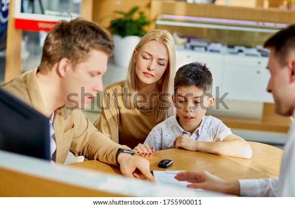 young caucasian family read documents and talk with\
consultant before purchase, they sit at table after view all cars\
represented there