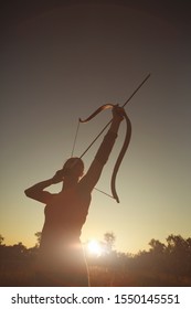 Young Caucasian female archer shooting with a bow in a field at sunset.