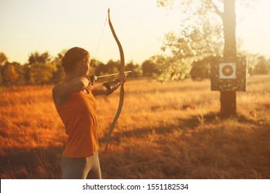 Young Caucasian female archer shooting with a bow in a field at sunset.