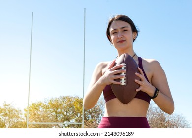 A young Caucasian female athlete holding an american football ball by the ropes. She is wearing an activity wristband and goal posts are visible in the background. - Powered by Shutterstock