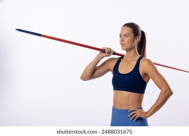 Young Caucasian female athlete holds a javelin on her shoulders, with copy space on a white background. She's in athletic wear, showcasing strength and focus in a studio setting. - Powered by Shutterstock