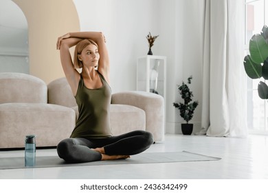 Young caucasian female athlete stretching her arms while doing yoga at home. Woman in sporty clothes relaxing in lotus position on fitness mat in the living room - Powered by Shutterstock