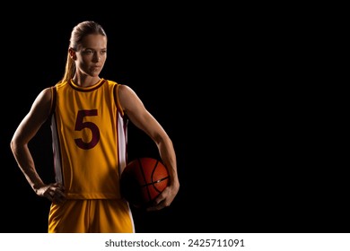 Young caucasian female basketball player poses in a basketball uniform on a black background, with copy space. Her stance and expression exude determination and athleticism on the court. - Powered by Shutterstock