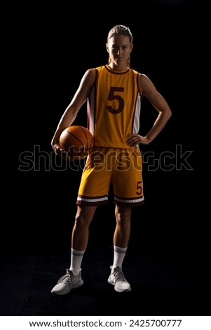 Young Caucasian female basketball player poses confidently in a basketball uniform on a black background. Her athletic stance and focused expression convey determination and skill.