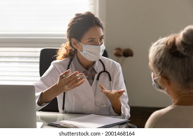 Young Caucasian Female Doctor In Facial Mask Talk Consult Old Patient In Hospital Or Clinic. Woman GP In Facemask Against Corona Covid-19 Have Consultation With Mature Client. Coronavirus Concept.