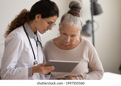 Young Caucasian Female Doctor And Old 60s Woman Patient Look At Tablet Screen Discuss Prescription Together. GP Or Therapist Consult Mature Client In Private Clinic Using Modern Pad Device.