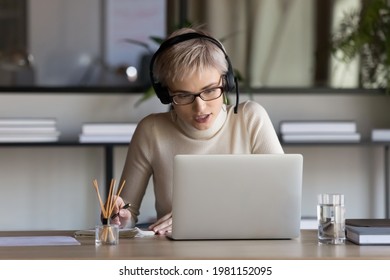 Young Caucasian female employee in earphones look at laptop screen talk speak on video call in office. Successful businesswoman in headphones have webcam online virtual conference on computer.