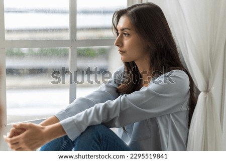 A young Caucasian female sits by the window at home on a rainy day, looking out with an absent-minded gaze. She appears sad and downcast, lost in her thoughts during the gloomy weather.