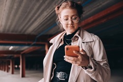 Young Caucasian Female Student, Generation Z, Dressed In A Trench Coat In The Parking Lot Uses A Smartphone