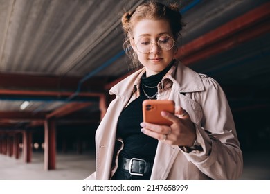 young caucasian female student, generation z, dressed in a trench coat in the parking lot uses a smartphone - Shutterstock ID 2147658999