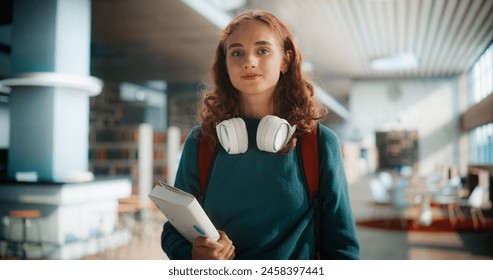 Young Caucasian Female Student With Headphones and Books Walking Through Modern Library. Confident Woman Carrying Backpack and Notebook, Ready for College Classes or Study Session. - Powered by Shutterstock