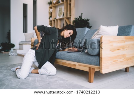 Young caucasian female suffer from back pain of remote job at home interior sitting near sofa, sad tired woman holding back feeling uncomfortable ache tension of spine arthritis chronic inflammation