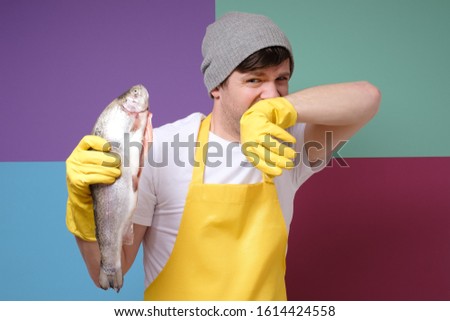 Young caucasian fisher holding a rotten fish isolated on colored background. Studio shot