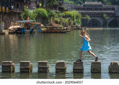Young Caucasian girl crossing waters on stepping stones on Tuojiang river, flowing through the centre of Fenghuang Old Town, China - Shutterstock ID 2029064057