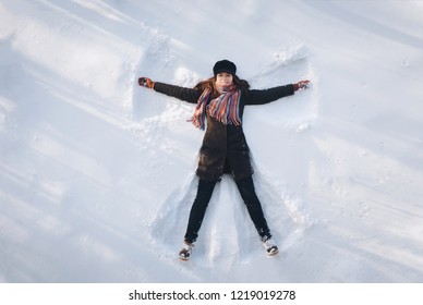 A Young Caucasian Girl On The Snow Shows An Angel. Spreading Arms. Top View. Drone. Smile. Happy.