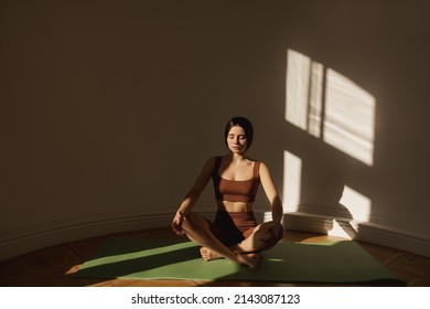 Young Caucasian Girl Practicing Yoga, Sitting On Mat In Relaxed Pose In Morning. Brunette Woman Exercising At Home Wearing Top And Leggings. Wellbeing, Mental Health Care Concept