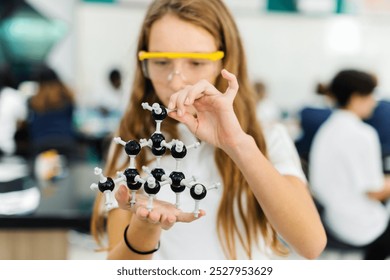 Young Caucasian girl in a science lab, holding a molecular model. The girl is focused on the molecular model, wearing safety goggles. Science lab, molecular model, girl. High school education. - Powered by Shutterstock