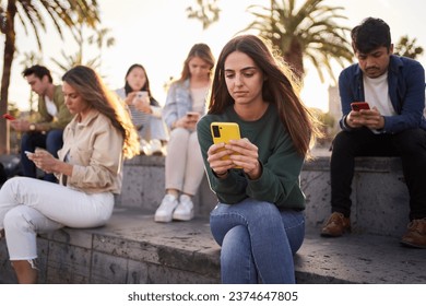 Young Caucasian girl sitting concentrated typing on mobile with group friends in background. Serious beautiful woman addicted to technologies using phone outdoors. Social isolation new generations. - Powered by Shutterstock