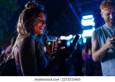 A young Caucasian girl is updating her social media with photos from a concert she's attending with friends holding beer in her hand and a smartphone. - Powered by Shutterstock