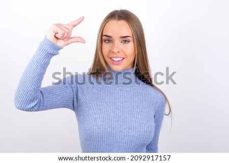 Young caucasian girl wearing blue turtleneck over white background smiling and gesturing with hand small size, measure symbol.