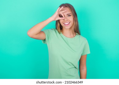 Young Caucasian Girl Wearing Green T-shirt Isolated Over Green Background Doing Ok Gesture With Hand Smiling, Eye Looking Through Fingers With Happy Face.