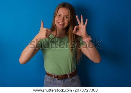 Young caucasian girl wearing green T-shirt over blue background smiling and looking happy, carefree and positive, gesturing victory or peace with one hand