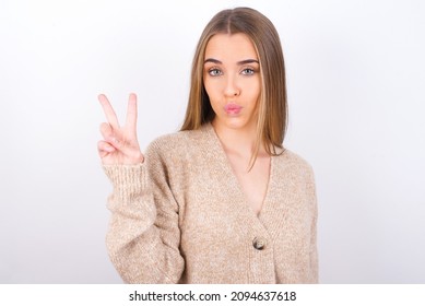 young caucasian girl wearing knitted sweater over white background makes peace gesture keeps lips folded shows v sign. Body language concept