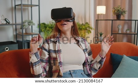 Young Caucasian girl wearing VR headset resting at home sitting on sofa in living room alone using futuristic technology making gestures with hands in air, play 3D video game. Virtual reality glasses