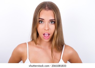 Young Caucasian girl wearing white tank top on white background having stunned and shocked look, with mouth open and jaw dropped exclaiming: Wow, I can't believe this. Surprise and shock