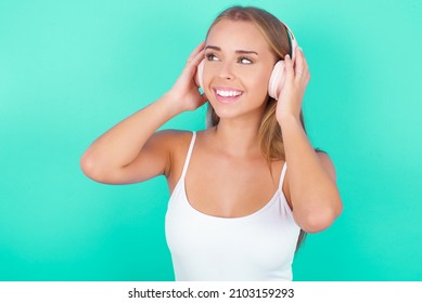 Young caucasian girl wearing white tank top over green background wears stereo headphones listens music concentrated aside. People hobby lifestyle concept