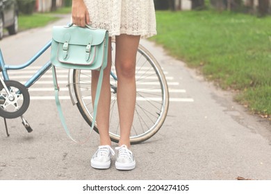 Young Caucasian Girl In A White Summer Dress, Bare Legs, Holding Mint Satchel Bag In Front Of The Bicycle On The Road