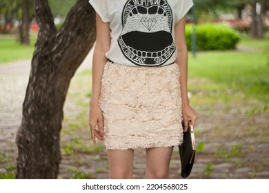 Young Caucasian Girl In A White T Shirt With A Print And Mini Pink Skirt With Textured Fabric Holding Black Clutch Bag In A Park