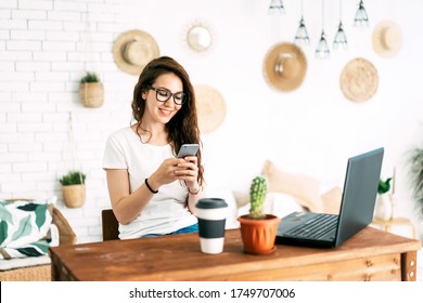 A Young Caucasian Girl At A Workplace In A Home Office Looks At A Smartphone. Working Businesswoman Girl Boss At Home.