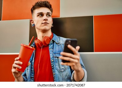 Young Caucasian Guy Drinking Soda Using Smartphone Leaning On The Wall